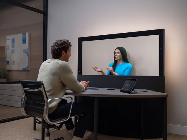 A seated person facing a remote participant on an HP Dimension display, with cameras in its frame and a meeting controller on the table.